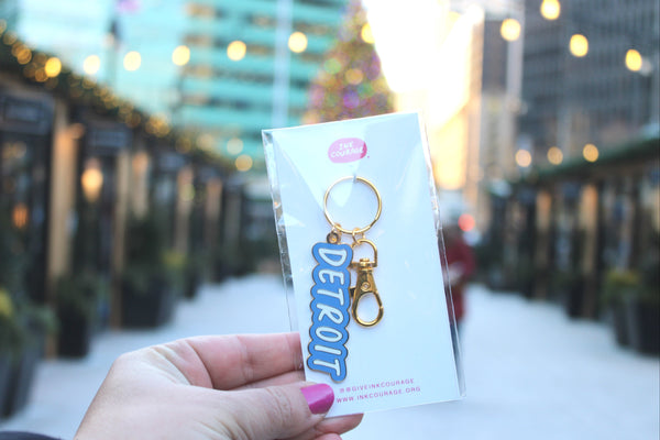 A hand with pink nail polish holds a packaged keychain featuring a gold-tone metal clasp and a blue die-cut charm that reads 'DETROIT' against a blurred outdoor city walkway background.